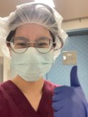 A young asian woman wearing hairnet, glasses, and blue face mask gestures a thumbs up with a gloved hand at the camera.